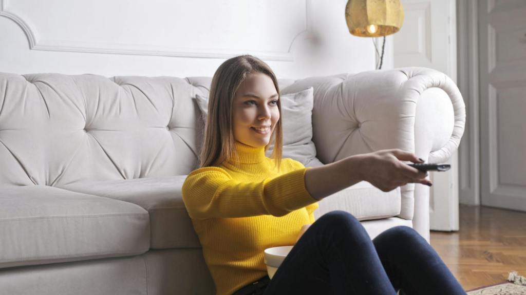 Vrouw in gele trui kijkt tv vanaf de zetel en stelt productieve taken uit – illustratie van present bias en uitstelgedrag