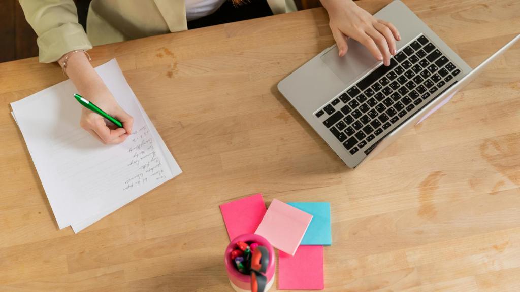 Vrouw schrijft op papier terwijl ze op haar laptop werkt, symbool voor het balanceren tussen denken en voelen bij het nemen van beslissingen – mental coach Wim Annerel.